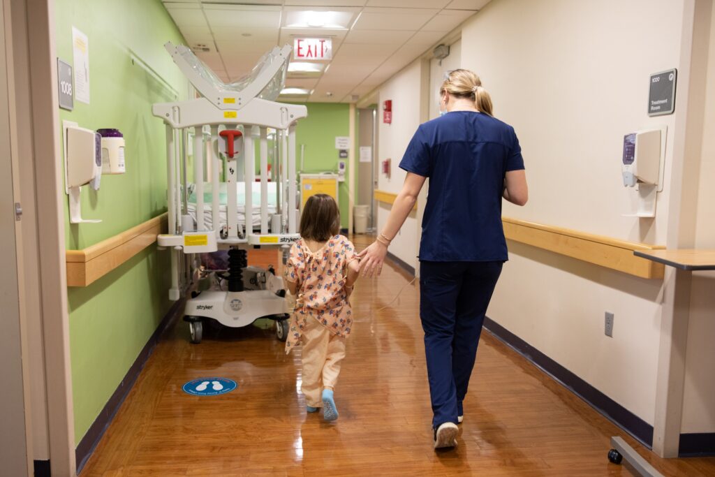 Mass Eye and Ear child life specialist Ashley Hoyt walks down the hallway of the pediatric floor at Mass Eye and Ear with a young patient.