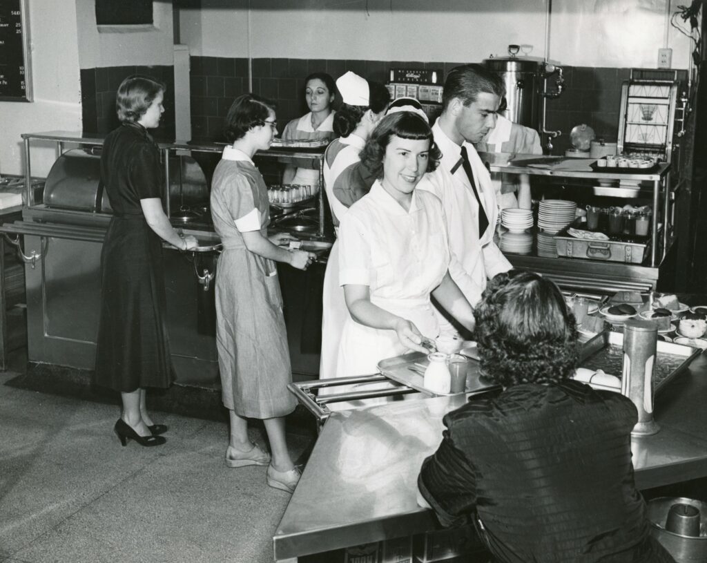 Black and white photo of several people getting food and checking out at the Mass Eye and Ear Cafeteria in 1952.