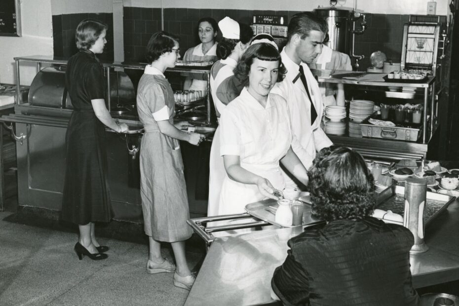 Black and white photo of several people getting food and checking out at the Mass Eye and Ear Cafeteria in 1952.